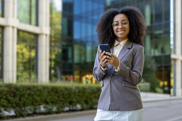 Confident african american business woman in gray blazer smiling, holding phone outdoors near modern office building. Concept of success, professionalism, and digital communication.