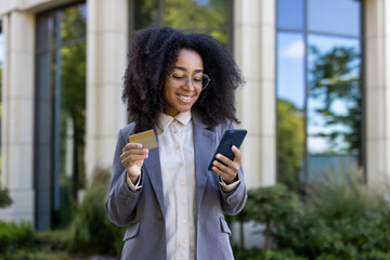 Confident african american business woman smiling with smartphone and credit card outdoors. Engaged in online shopping or banking. Casual office attire, glasses, natural hair.