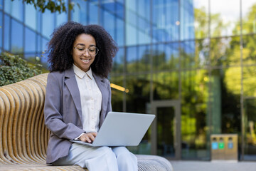 Confident African American businesswoman seated on stylish wooden bench using laptop outdoors, demonstrating modern work-from-anywhere lifestyle. Smiling professional symbolizes empowerment and