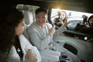 Medium shot of smiling male celebrity in classy grey suit talking to unrecognizable woma sitting in...