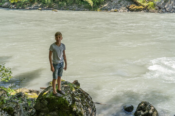 A young boy stands on a large rock by the river, surrounded by greenery, observing the flowing water on a bright day