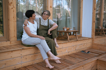 Naklejka premium Two women talk, hugging each other while sitting on terrace of country house on summer day