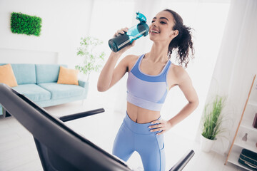 Joyful woman hydrating during home treadmill workout in stylish sportswear