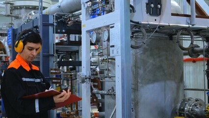 Industrial worker in safety gear checks machinery in factory, pointing at pressure gauge with clipboard. Industrial worker holding clipboard and pointing at pressure gauge while inspecting machinery