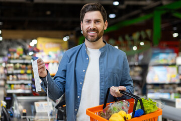 Smiling man at grocery store checkout holding receipt and orange shopping basket filled with fresh vegetables. Expresses feeling of satisfaction and successful shopping experience.