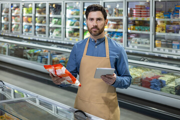 Store employee in apron holds digital tablet and product in frozen food section of grocery store,...