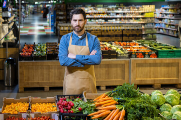 Grocery store worker in apron stands confidently with arms crossed. Surrounded by fresh produce like carrots and tomatoes in modern supermarket. Brightly lit store interior.