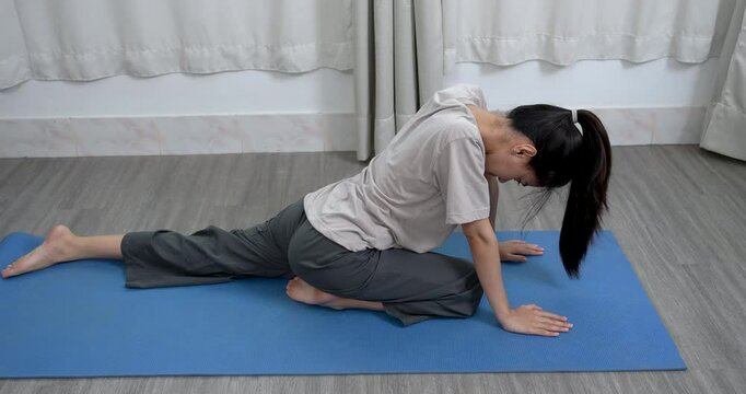 Asian woman stretching forward in yoga split pose on blue mat indoors. Calm expression, stretching hamstrings and back, peaceful home environment with plant and curtains.