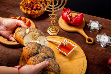 Female hands breaking challah on the table in Rosh Hashanah next to an apple, pomegranate, honey, menorah
