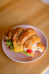 Сroissant sandwich on a pink plate, placed on a wooden table. The croissant is filled with lettuce, a slice of cheese, mayonnaise, and bright red peppers, creating a colorful and appetizing look. 