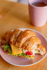 Сroissant sandwich filled with cheese, lettuce, and vegetables, served on a pink plate. Behind it, there is a matching pink cup, creating a soft and cozy atmosphere with a warm, blurred background.