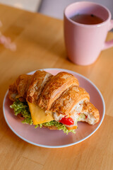 Сroissant sandwich filled with cheese, lettuce, and vegetables, served on a pink plate. Behind it, there is a matching pink cup, creating a soft and cozy atmosphere with a warm, blurred background.