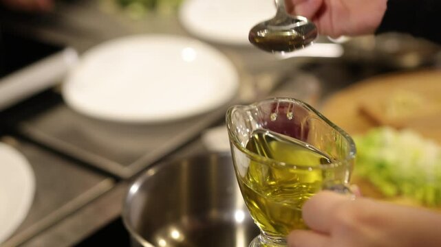Kitchen setting, cooking oil, kitchen prep. Hands drizzling spoonful olive oil into bowl in kitchen setting, with background out of focus.