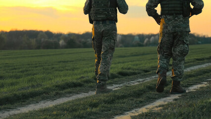 Soldiers walking through field at sunset. Two soldiers in camouflage uniforms walk through a grassy...
