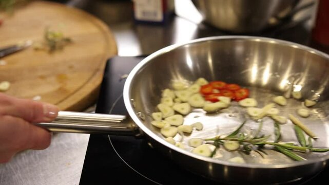 Chili peppers, frying spices, garlic cooking. Close-up image captures garlic slices, chili peppers, rosemary sprigs sizzling in oil inside stainless steel pan.