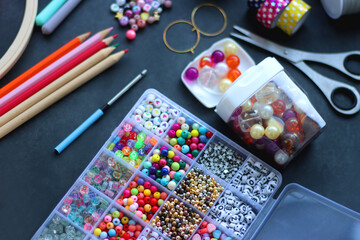 Colorful beads, coloring pencils, glitter, needles, string, hoop, tape and scissors on dark background. Various colorful craft supplies. Selective focus.