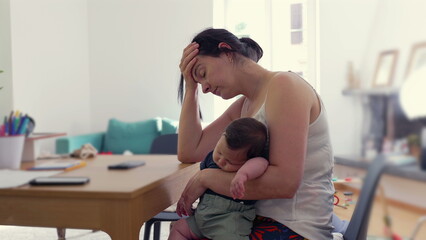 Mother sits at the table, head in hand, cradling a sleeping baby, embodying the fatigue and stress of motherhood, in a moment of quiet overwhelm in a home environment