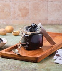 Green walnut jam in a glass jar on a wooden tray on a green concrete background. Exotic sweets, recipes from around the world.