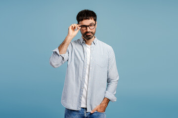 Fototapeta premium Handsome, serious, bearded man wearing stylish eyeglasses, standing isolated on blue background