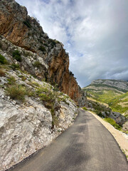 Winding road cuts through a steep mountain landscape, framed by towering rock formations and a moody sky. Harmony between man-made paths and the grandeur of untouched nature