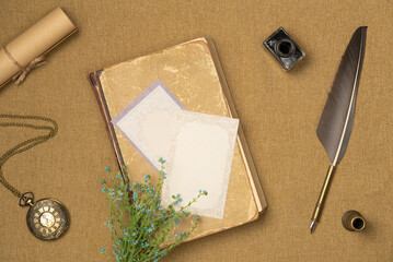 Paper blank sheet on book with ink, quill pen, watch and flowers on fabric background from above. Vintage style. Flat lay, top view.