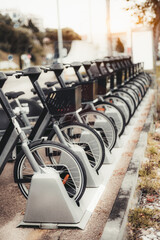 A vertical shot of a row of parked rental bicycles, each secured in a docking station on an urban street. The bicycles are equipped with front baskets and sleek designs, ready for public use