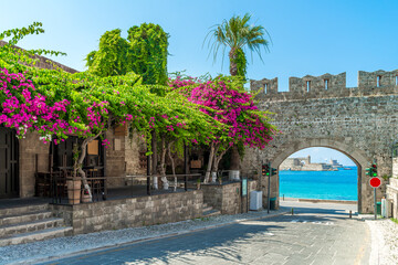 Picturesque Virgin Mary Gate in Rhodes Old Town with a medieval stone arch, vibrant bougainvillea, and a stunning coastal view of the Mediterranean Sea.