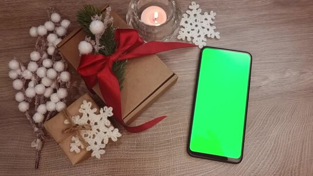 Green screen smartphone lying on wooden table next to burning candle, gifts fnd DIY Christmas decorations. Christmas mockup top view.