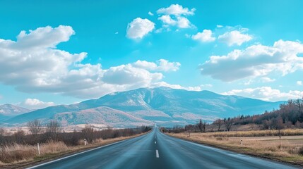 A straight road leads towards a mountain range under a clear blue sky with white clouds.