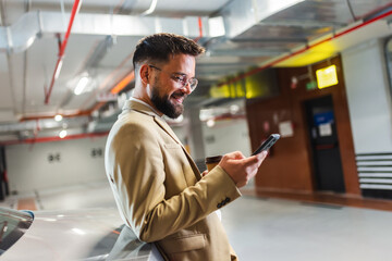 Portrait of businessman using smartphone and drinks coffee while standing near his car in...