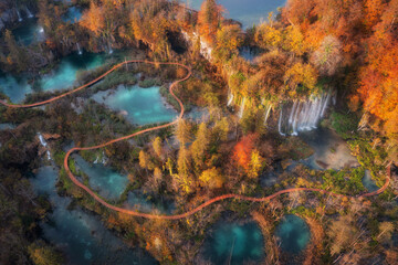 Aerial view of waterfall in red autumn forest at sunrise. Morning in Plitvice Lakes park, Croatia. Colorful landscape with waterfall, trees, wooden trails and bridges, orange foliage, river. Top view