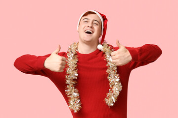 Fototapeta premium Handsome young man in warm sweater, Santa hat and tinsel showing thumbs-up on pink background