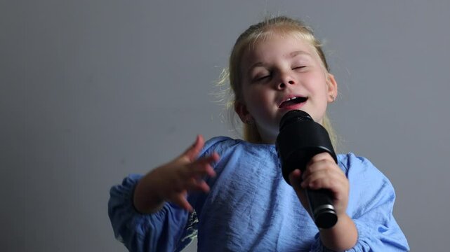 A little girl sings while holding a microphone. Young beautiful actress with blonde straight hair and a blue suit. Karaoke for children, young singer