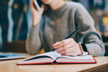 Selective focus on female hand making notes in textbook for education while calling to operator for consultancy, woman preparing to exams using notepad during e learning with laptop computer