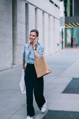 Stylish smiling hipster girl dressed in branded wear calling on smartphone device and talking with friend using high speed connection walking outdoors around street, positive shopper with bags