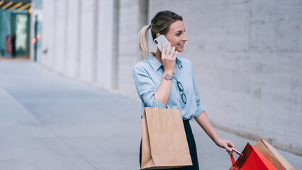 Cheerful young woman dressed bought branded clothes on sales during black friday, happy girl walking on street with paper bags while communicate via smartphone device using 4G wireless internet