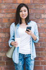 Full length portrait of attractive Chinese hipster blogger with caffeine beverage in hand posing with cellphone on brick background, millennial woman dressed in casual denim apparel enjoying trip time