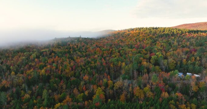 Aerial view of a colorful autumn forest covered in fall foliage, with vibrant reds, oranges, and yellows. Mist rises over the treetops as the morning sunlight illuminates the landscape.