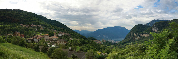 Fototapeta premium Panorama Aussicht ins Tal mit mehreren mediteranen Italienischen Bergdörfern, Wald und einem See im Sommer bei bewölktem Himmel.