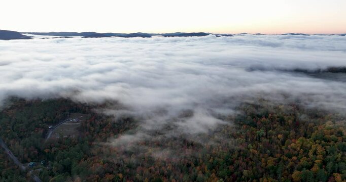 Aerial view of a mountain range covered in vibrant fall foliage, with mist and fog drifting over the colorful forested hills, capturing the beauty of the autumn season.
