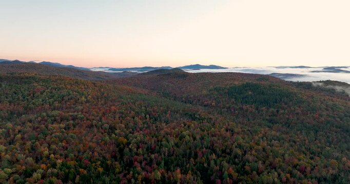 Aerial view of a mountain range covered in vibrant fall foliage, with mist and fog drifting over the colorful forested hills, capturing the beauty of the autumn season.