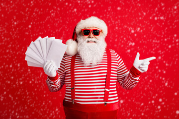 Photo of grandfather grey beard direct finger empty space hold fan envelopes wear santa claus x-mas costume suspenders sunglass striped shirt cap gloves isolated red color background