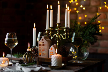 Luxury elegant Christmas dinner table setting with old vintage golden chandelier, candles, fir tree branches, wooden furniture. Porcelain houses as centrepiece, garland with lights on background 