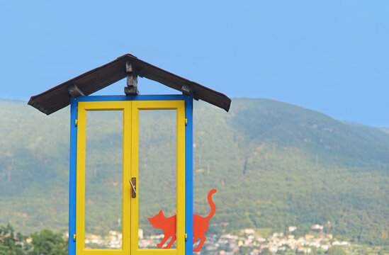 Fake red cat in bright yellow and blue window welcoming visitors in Brolo, the cat village at Lake Orta, with mountains in the back and blue sky on a summer day. Selective focus
