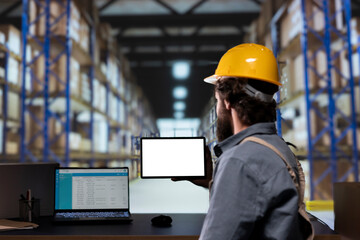 Warehouse manager looking at display with white layout on tablet, working in storage room depot...