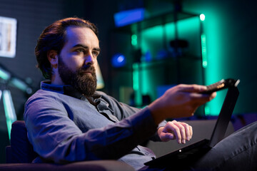 Man sitting at home on couch working on laptop while picking TV channel to watch as distraction. Remote employee solving company tasks on notebook and browsing movies using remote