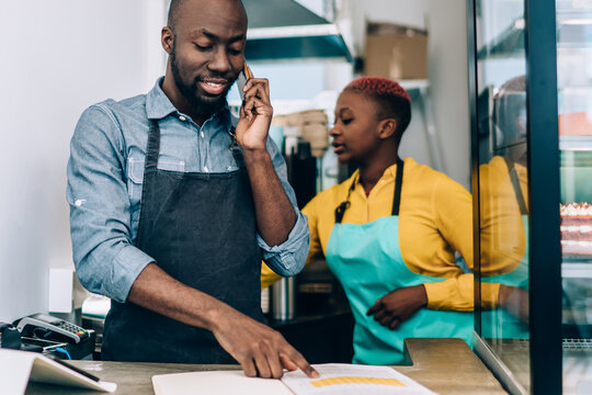 Black waiter checking data in notebook while talking on phone