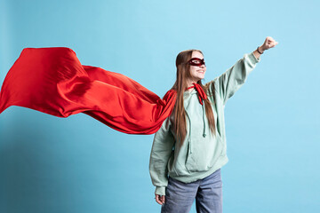 Cheerful girl wearing superhero costume and mask for Halloween with cape fluttering, studio background. Portrait of jolly teenager smiling while dressed as comic book hero, pretending to fly © DC Studio