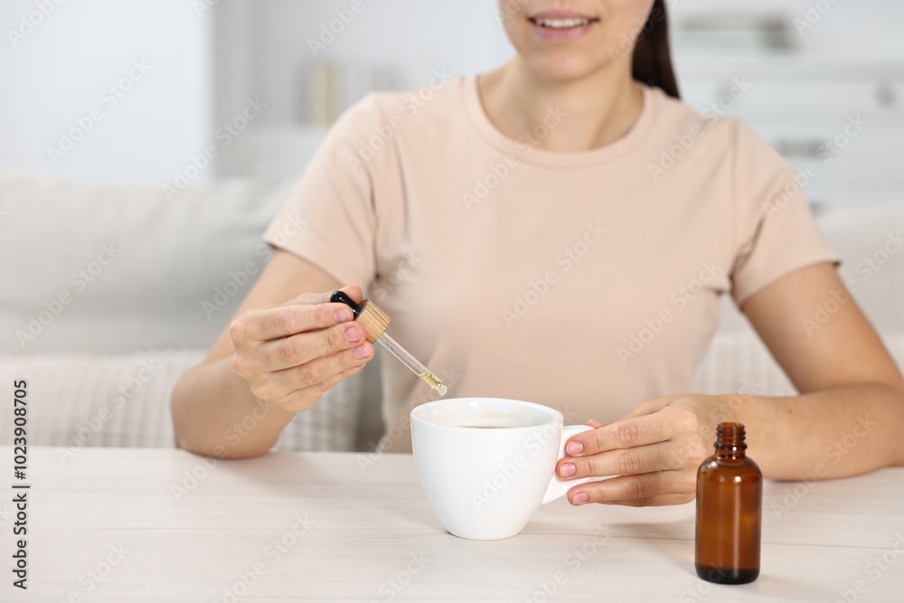 Canvas Prints Young woman putting CBD tincture into cup with drink at white wooden table, closeup