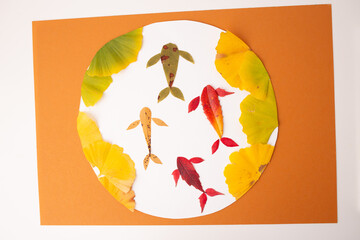 Autumn leaf art depicting fish arranged in a circular pattern with yellow ginkgo leaves on a white and orange background. Flat lay composition. Nature seasonal craft concept.